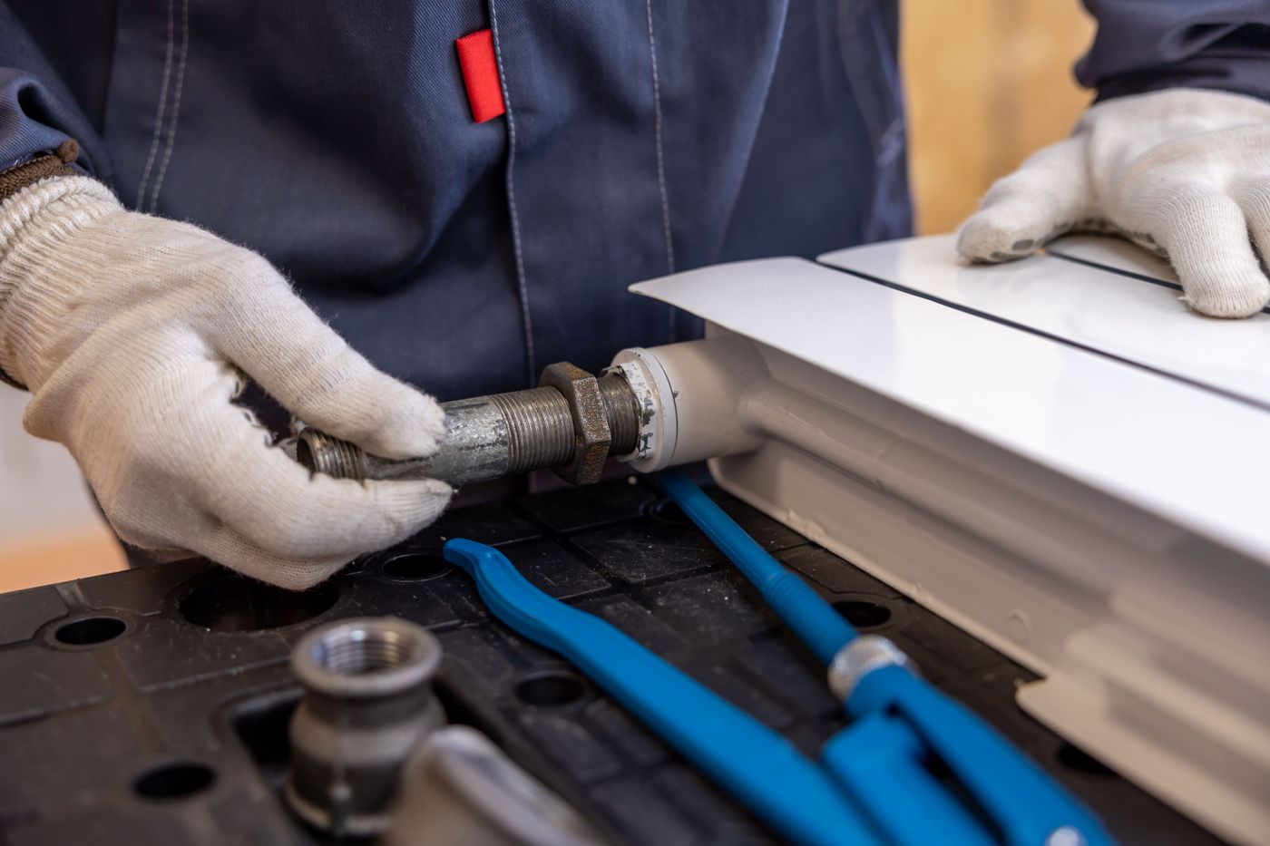 Gloved hands fitting a threaded pipe connection, with plumbing tools laid out below.