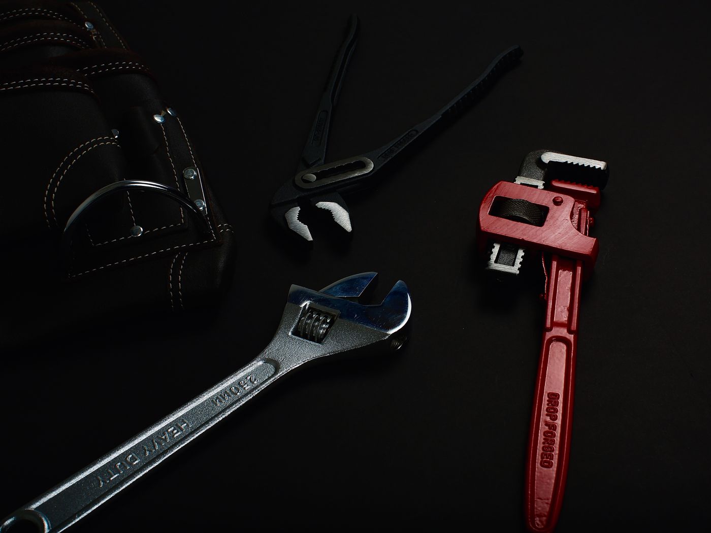 A pipe wrench, adjustable spanner and water pump pliers on a dark surface.